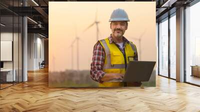 Portrait of power engineer wearing safety jacket and hardhat with laptop computer working at outdoor field site that have wind turbine at the background. Wall mural