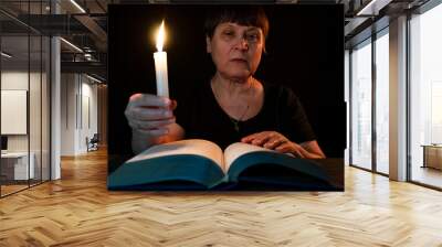 An elderly woman is reading a book in the dark with a burning candle. Wall mural