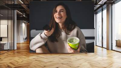 Brunette woman eat bucket of pistachio ice cream and watch tv, change channel with remote control sitting on bed in bedroom. Happy laughing girl watch comedy movie, have fun at home. Wall mural