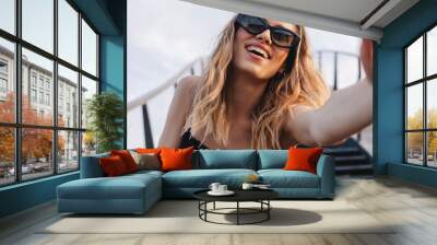 Cheerful lady making selfie at sunny day. Pretty young girl with curly blonde hair and two thin braids towards to camera, smiling and wear black glasses and dress under the stairs bridge and sky. Wall mural