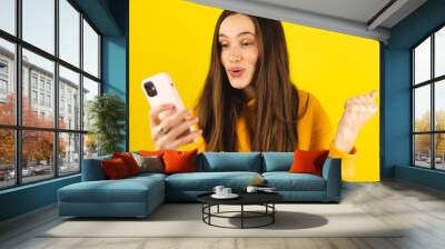 Fun young woman hold in hand use mobile cell phone doing winner gesture clenching fists isolated on yellow background studio. Girl use smartphone apps winning online celebrating discount gift voucher. Wall mural