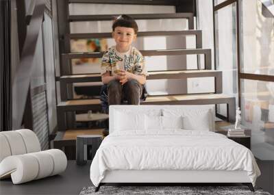 Happy little boy sitting on the stairs in cafe. Cute boy kid look at side with fun hair cut. Wall mural