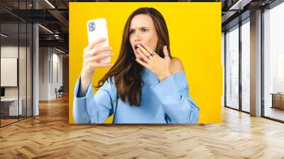 Mad and shock young brunette beautiful woman using mobile phone isolated on yellow background. Yelling unhappy woman scream cover mouth. Disappointed sad upset lady horrified impressed news. WTF Oh no Wall mural