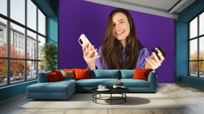 Online shopping. Portrait of smiling brunette hair woman paying with plastic credit card on smartphone app, standing with mobile phone and bank card against purple background. Happy emotions. Wall mural