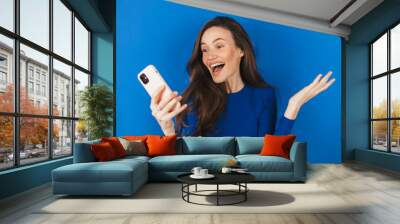 Overjoyed happy excited woman say wow, glad to receive text message informing about salary, rejoices good news, stares at mobile phone, gestures actively from happiness isolated blue background. Wall mural