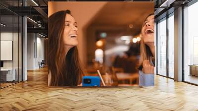 Overjoyed two women friends holding mobile phone shopping online with sales, win money, celebration success sitting in modern cafe. Happy successful freelancers receive payment, reading good news. Wall mural