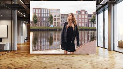 Pensive blonde woman wear black dress, leather jacket and touch it, posing, turn around. Outdoor shot of calm hippie lady with two thin braids and wave hair. Coachella or boho freedom style. Wall mural