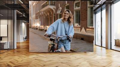 Portrait of a pretty young woman on bicycle in the city street, city transportation. Outdoor fashion portrait of elegant blonde curly lady riding her hipster retro bike in stylish blue warm sweater. Wall mural