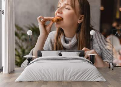 Young blonde woman with bang eating croissants at a cafe. Girl bite piece of croissant look joyful at restaurant. Cheat meal day concept. Woman is preparing with appetite to eat croissant. Wall mural