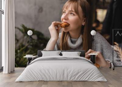 Young blonde woman with bang eating croissants at a cafe. Girl bite piece of croissant look joyful at restaurant. Cheat meal day concept. Woman is preparing with appetite to eat croissant. Wall mural