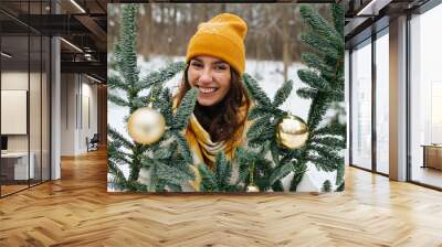 Happy smiling girl outside at winter behind synthetic fir tree decorated with shiny christmas balls. She wears yellow knit scarf and hat. Wall mural