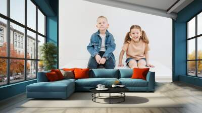 Little siblings sitting on a table, posing for a photo. Boy is sitting on his knees, looking with subtle smile on his face. Cute girl with pony tails is leaning on her hands, also smiling. Wall mural