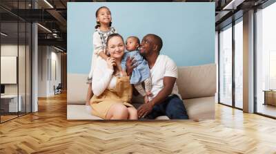 Portrait of happy multiethnic family sitting on couch at home and looking at camera. Black father, white mother and two daughters. Eldest leaning on mother from behind. Youngest is in father's arms. Wall mural