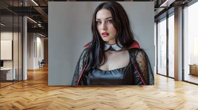 Young woman dressed as a vampire in a sleek black and red costume, Halloween themed web design or digital marketing. Soft, neutral background enhances the dark tones of the outfit Wall mural