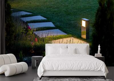 marble path of square tiles illuminated by a lantern made of metal glowing with a warm light in a backyard garden with a flower bed and a lawn. Wall mural