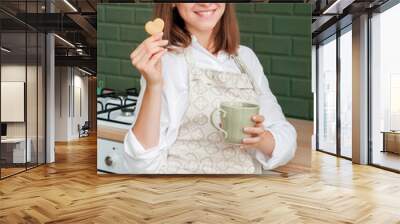 Happy slender young woman in an apron with a mug and cookies is sitting at home in the kitchen. Young hostess in the kitchen. Cooking cookies at home. Pastry chef girl Wall mural