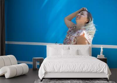 A beautiful woman straightens her hair against a blue wall on a sunny summer day. Portrait, copy space. On a sunny day, a smiling woman poses against a bright blue wall. Summer portrait concept. Wall mural