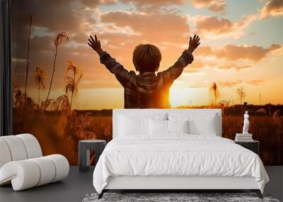 A little boy raises his hands above the sunset sky, enjoying life and nature. Happy kid on a summer field looking at the sun. Silhouette of a male child in the sun. Fresh air, environment concept. Wall mural