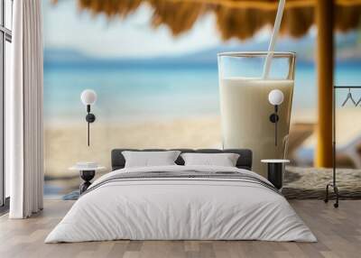 A glass of white milk with a straw in it is on a table next to a beach umbrella Wall mural