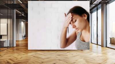 A close-up portrait of a young woman standing sad and holding his head in his hand and a white brick wall in the background. Headache and worries about illness, unemployment, temperature. Copyspace Wall mural