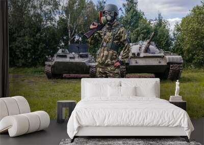 A soldier in a gas mask with a Kalashnikov rifle against the background of armored vehicles. Wall mural