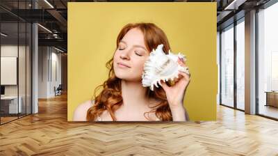 A young woman put a sea shell to her ear. The girl with red hair closed her eyes and listens to the sound of the sea in the sea shell, standing against the wall on a yellow background. Wall mural