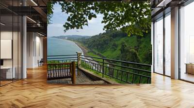 An observation deck with a bamboo bench in the shade of deciduous trees. View of the blue sea and the coastline to the horizon with a high, green-covered shore. Batumi Botanical Garden Wall mural