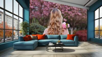 Beautiful young woman with long curly blonde hair from behind holding blooming branch of sakura tree Wall mural