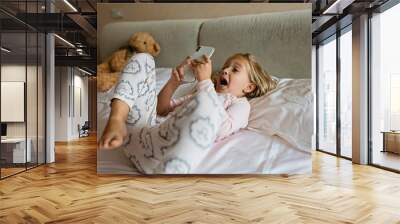 Cute little kid girl watching video on smartphone with smiley face alone on the bed, child using mobile phone with happy face at home. Stay at home quarantine coronavirus COVID-19 pandemic prevention. Wall mural