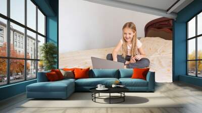 Stay at home quarantine coronavirus pandemic prevention. Little girl with blonde hair sitting on the bed and using laptop. Prevention epidemic. Child using technology. Distance online education Wall mural