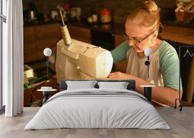 Confident senior elderly woman seamstress white shirt and glasses sitting in front of sewing machine, working on clothes at home using beige fabric. Retired people, age, job occupation concept Wall mural