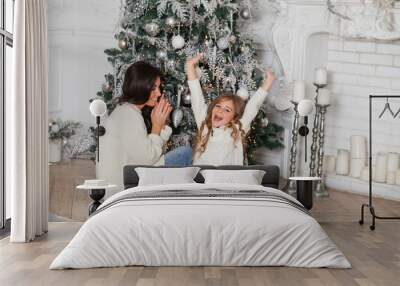 Happy family on New Year Eve. Young beautiful mother and little cute daughter in white cozy sweaters and blanket have fun and hug under a decorated Christmas tree. Festive home classic interior Wall mural