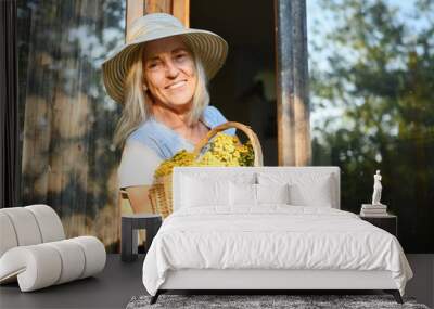 Happy smiling elderly woman having fun posing by open window in rustic old wooden village house in straw hat with flower basket. Retired old age people concept. Quarantine in the country house. Wall mural