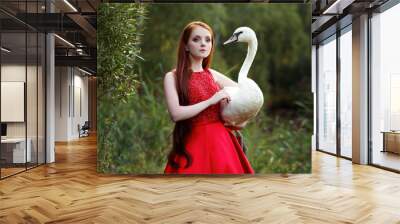 Beautiful long haired girl in red evening dress holding white swan in hands in the forest. Fairytale portrait of red head young woman Wall mural