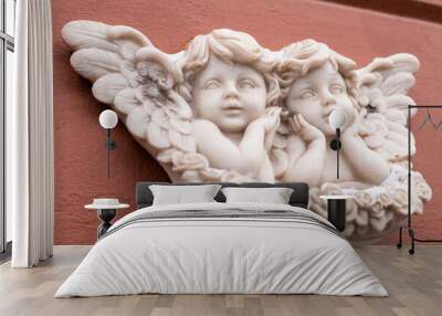 Two baby angels with wings marble sculpture on red wall of building. Close-up, front view. Wall mural