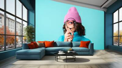 Cozy portrait of a young woman in a knitted blue sweater and a pink hat with bright makeup holding a magnifying glass, fooling around, having fun Wall mural