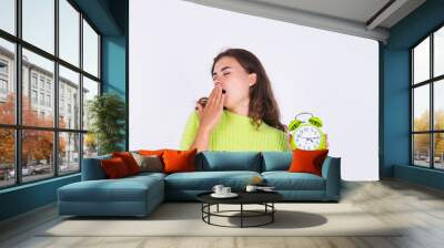 Young beautiful woman with freckles light makeup in sweater on white background with alarm clock angry tired sleepy exhausted yawning Wall mural