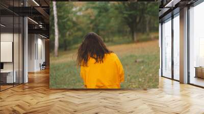 A woman in a bright yellow top walks through a park, smiling happily as she enjoys the beauty of nature and the warmth of the sun in autumn. Wall mural