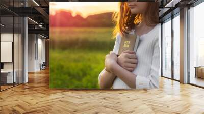 Christian teenage girl holds bible in her hands. Reading the Holy Bible in a field during beautiful sunset. Concept for faith, spirituality and religion. Peace, hope Wall mural