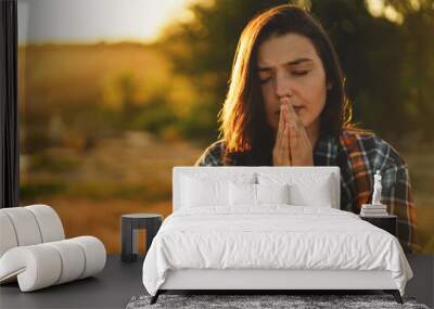 young woman closed her eyes, praying in a field during beautiful sunset. hands folded in prayer conc Wall mural