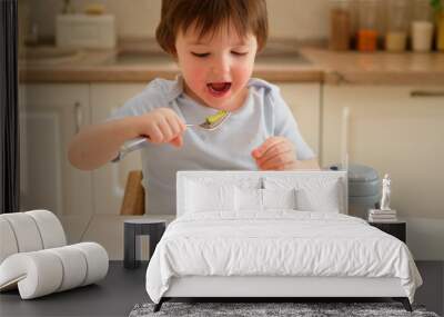 A child eats vegetable salad with a fork while sitting at a table in a home kitchen. Little boy eating in the kitchen at home. Toddler baby two years old at dinner Wall mural