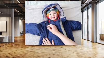 A mother woman puts on a warm jacket for a baby boy. Mom dresses a happy child in winter clothes on the bed. Wall mural