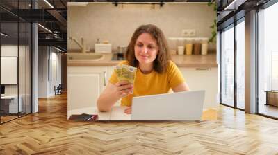 A woman with a laptop holds euro money at a table in a home kitchen. An adult female businesswoman works from home, a remote office Wall mural