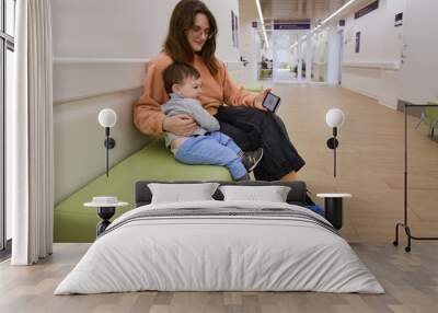 Mother and baby with a mobile phone in the clinic waiting for a pediatrician. Parent with a child boy with a smartphone in his hands. Kid is a boy aged one year and three months Wall mural