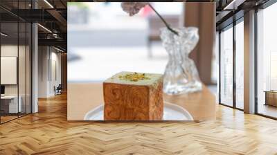 Close-up side view of freshly baked brown cube croissant with green pistachio cream on wooden table by flower in vase in restaurant or cafe. Soft focus. Copy space. Puff pastry theme. Wall mural