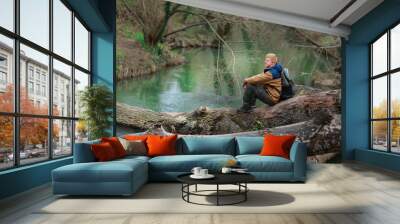 Redhead man in a brown-and-blue windbreaker with a backpack sits on a log by the green river in the spring in the Crimea. Wall mural