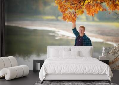 Redhead man in a plaid shirt holding an oak branch with orange leaves by the lake in the autumn forest Wall mural