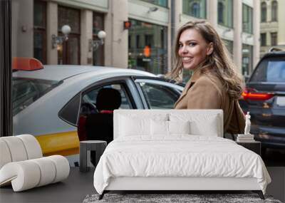 A beautiful young woman gets into a yellow taxi in the city. Smiling blonde in a beige coat. Travel and traffic in the metropolis. Wall mural