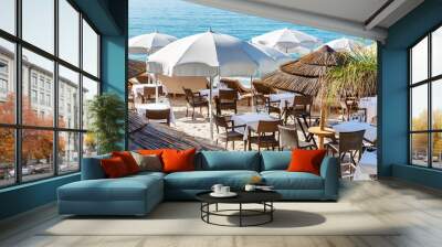 Restaurant tables on a sandy beach under thatched roof umbrellas. Relax and service at a fashionable resort. Wall mural