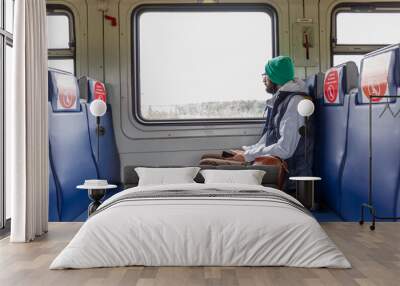 Stylish young man in glasses sits in a train car with markings for seating passengers and looks out the window. Wall mural